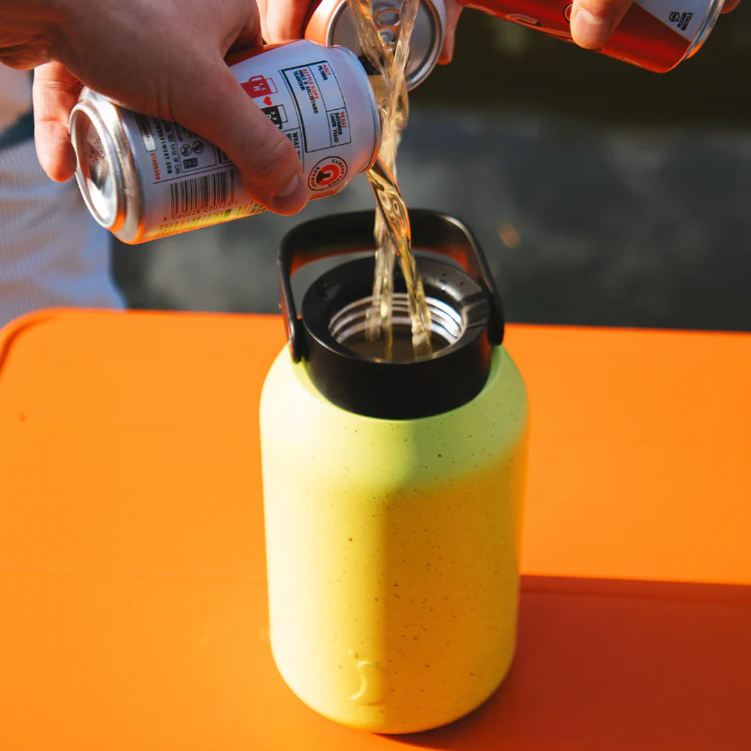 Person pouring beer from a can into a green bottle with a black cap on an orange surface.