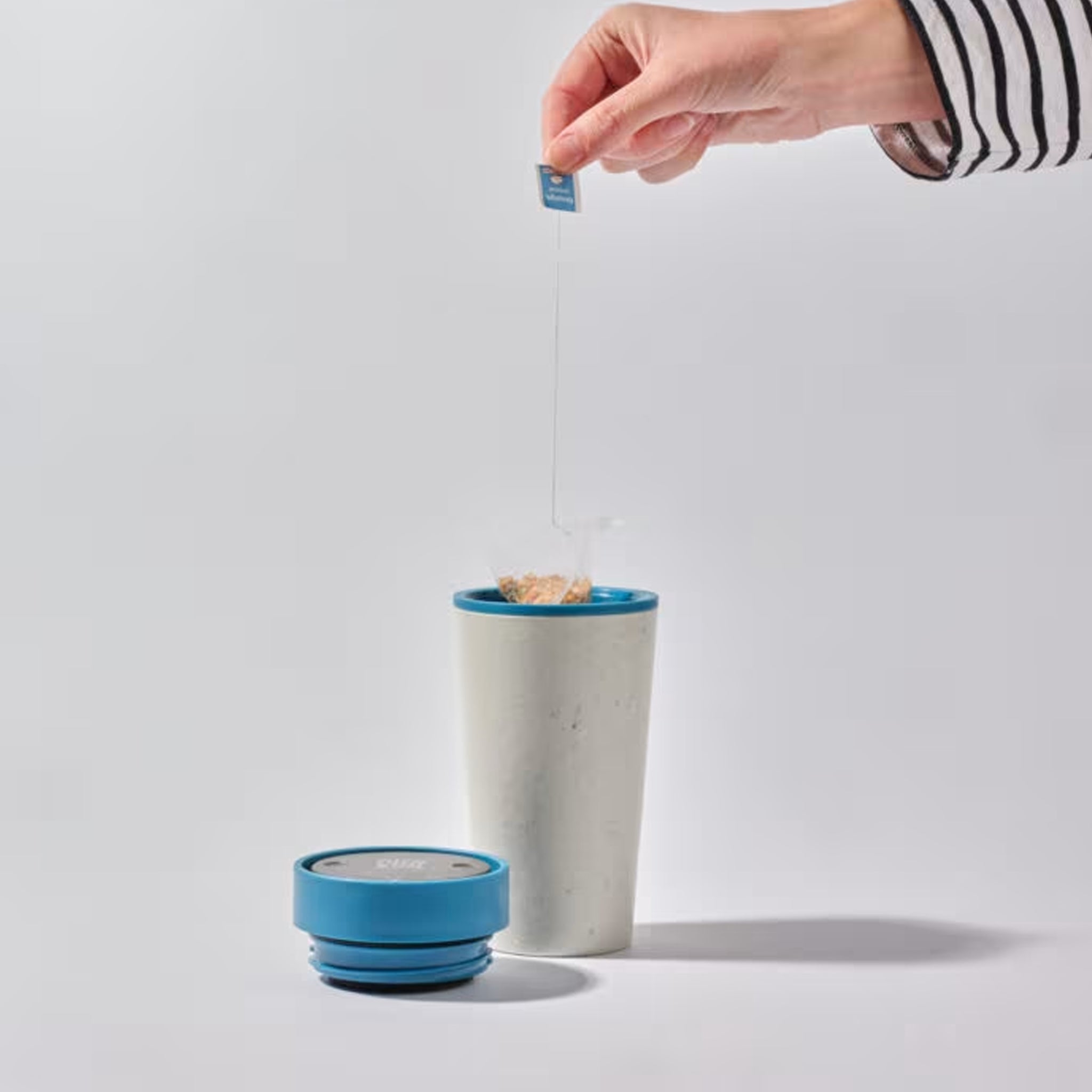 Person holding a tea bag over a white mug with a blue lid on a light gray background