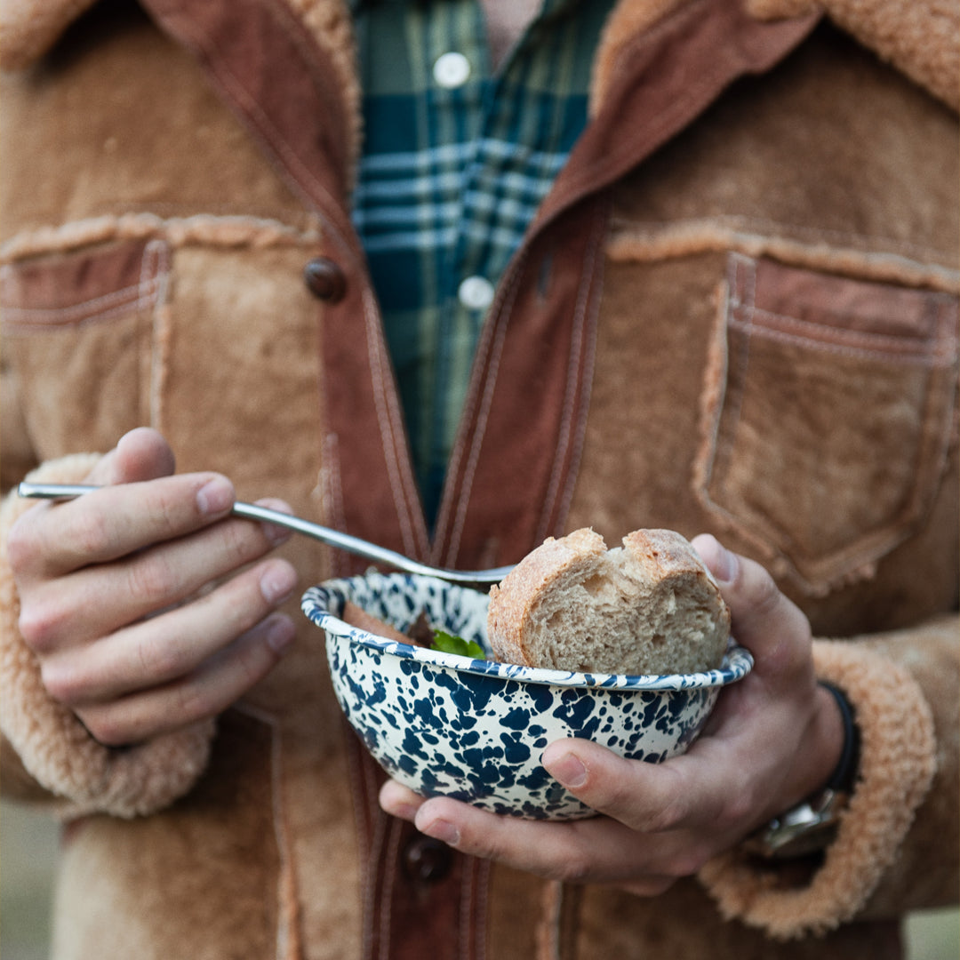 Person holding a bowl of soup with bread and a spoon, wearing a brown jacket and plaid shirt.
