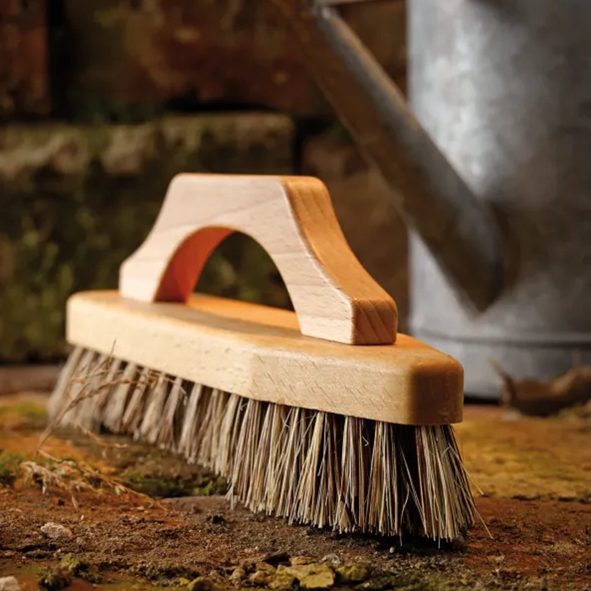 A wooden garden brush with natural straw-colored bristles, shown resting on wet tiles outdoors with a watering can blurred in the background.