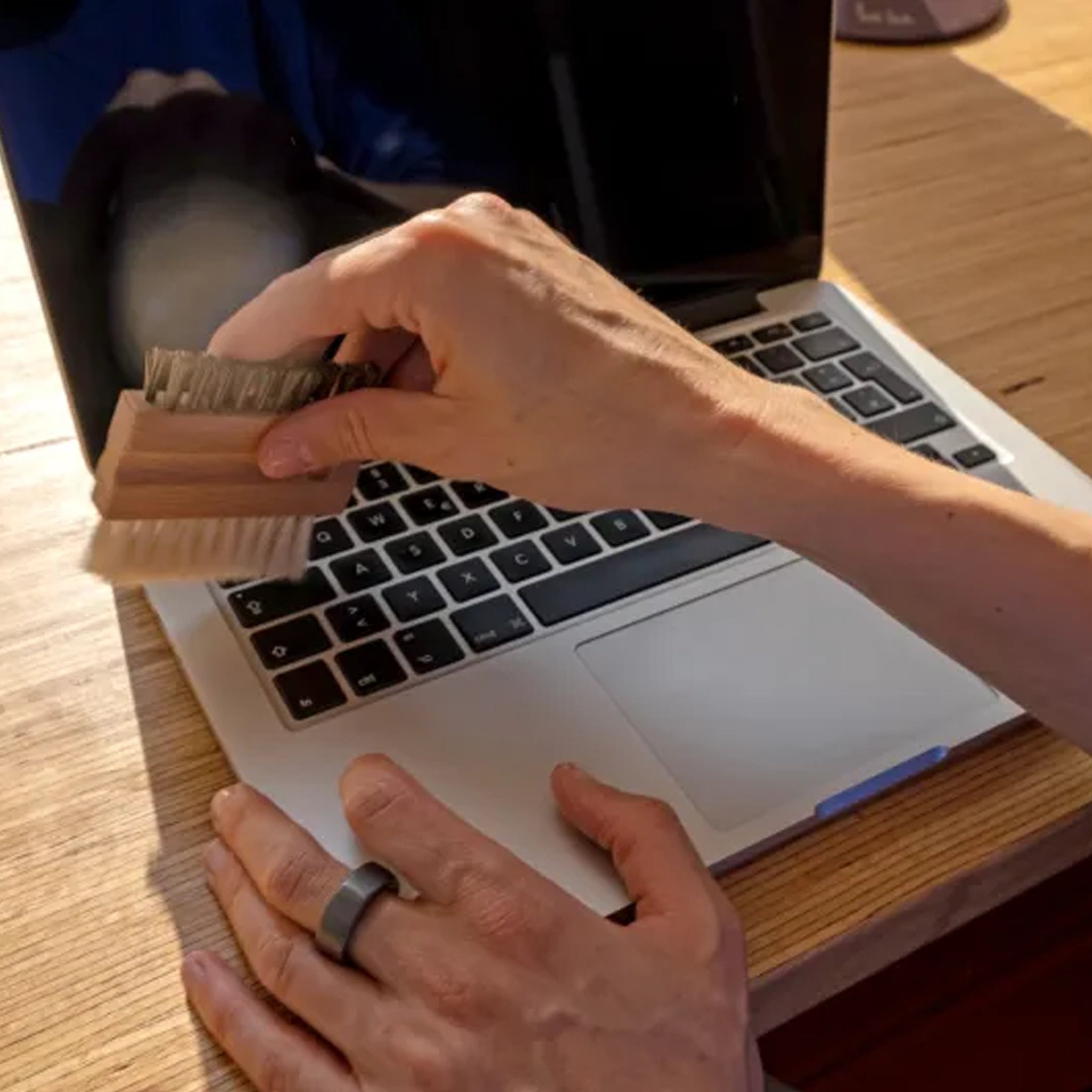 Person cleaning a laptop keyboard with a brush on a wooden desk.