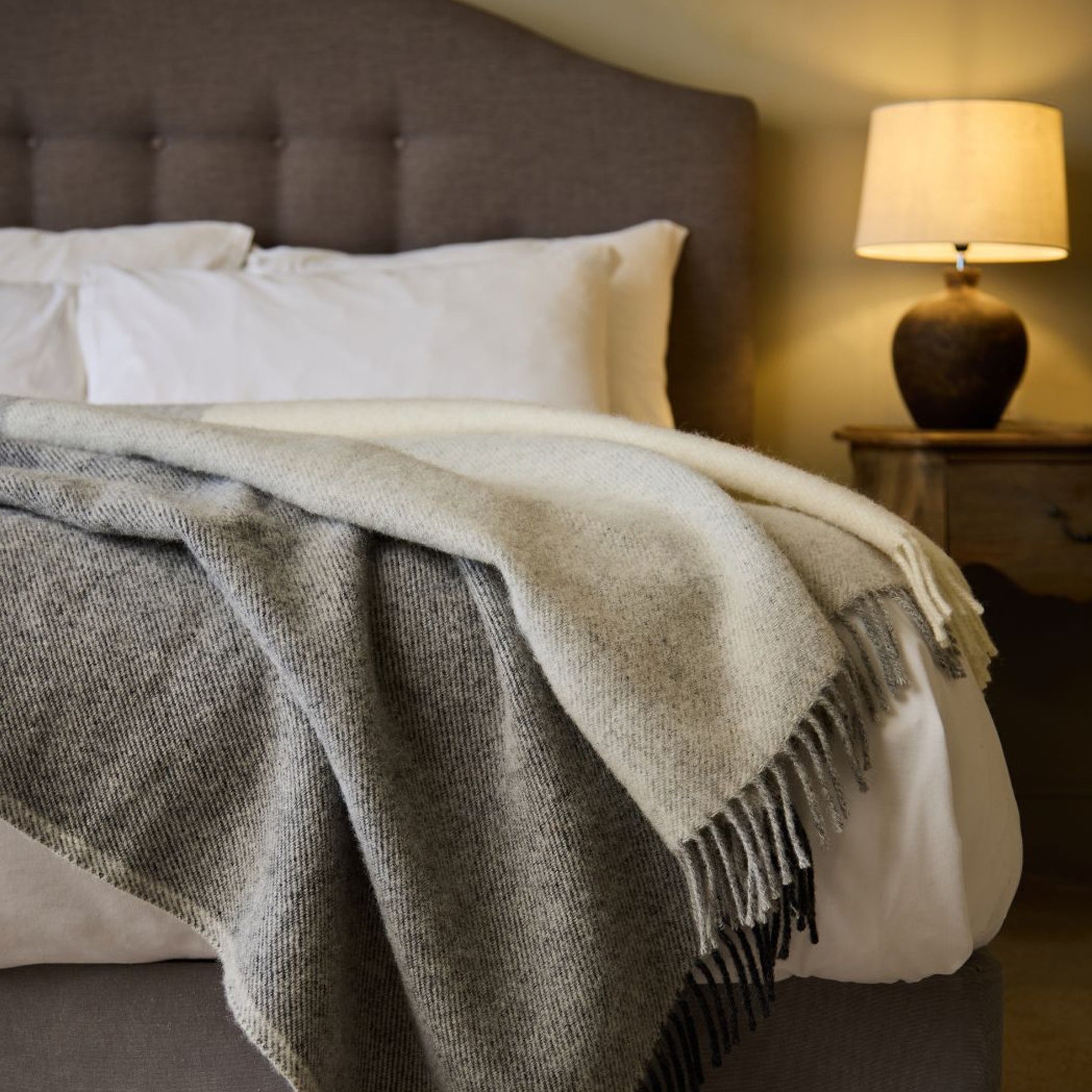 Bed with gray and white blanket, white pillows, and a lamp on a nightstand.