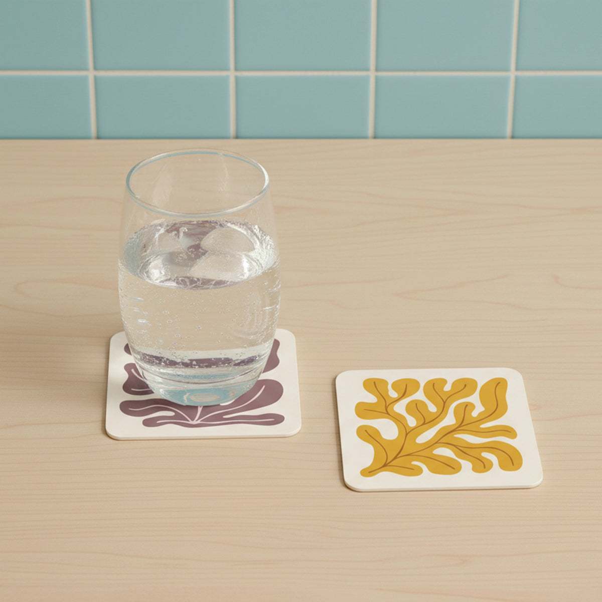 Two square coasters on a light wood tabletop with a pale blue tiled backsplash. One coaster is white with a dark purple abstract leaf-like pattern, and the other is white with a mustard yellow abstract leaf-like pattern.