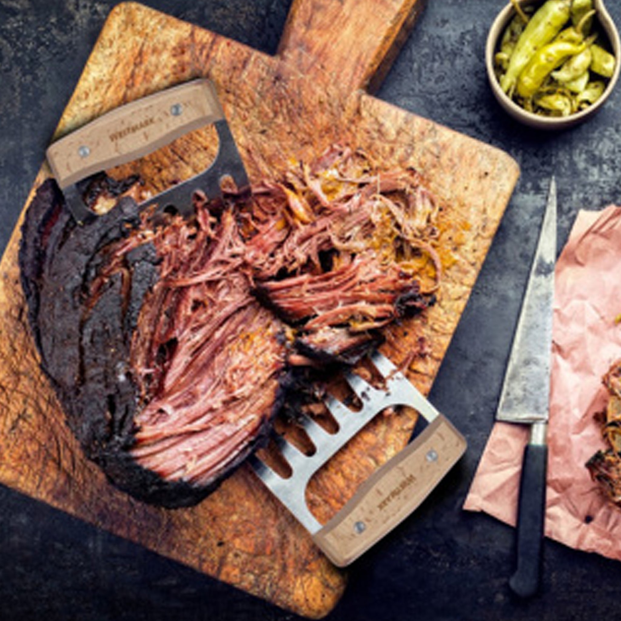 Shredded beef on a wooden cutting board with a meat claws, knife, and bowl of chillis.